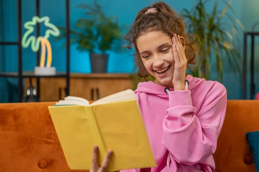 Caucasian preteen school girl reading funny interesting fairytale story book, leisure hobby, knowledge wisdom, education, learning, study, wow. Young child, kid at home room apartment sitting on couch