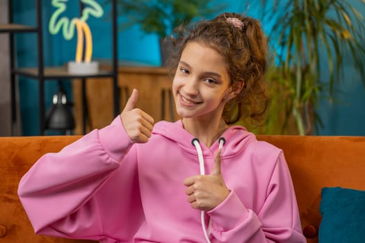 Like. Happy Caucasian preteen school girl looking approvingly at camera showing thumbs up, like sign positive something good, positive feedback. Young child kid at home room apartment sitting on couch