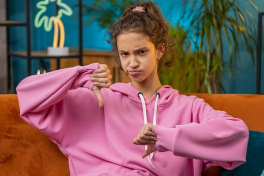 Dislike. Upset Caucasian preteen school girl showing thumbs down sign gesture, expressing discontent, disapproval, dissatisfied bad work. Displeased child kid at home room apartment sitting on couch