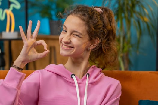 Okay. Happy cheerful preteen school girl looking approvingly at camera showing ok gesture, positive like sign, approve something good, winking, blinking her eye. Child kid at home room on couch