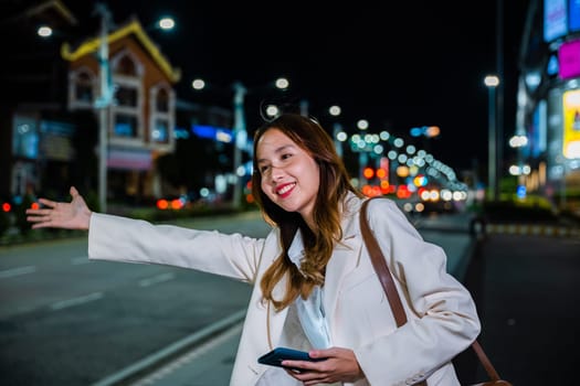 Asian businesswoman standing hail waving hand taxi on road in busy city street at night, beautiful woman smiling using mobile phone application hailing with hand up calling cab after late work