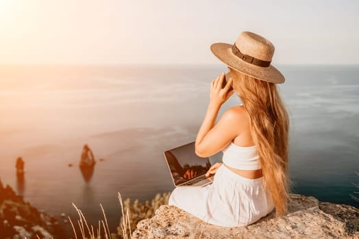 Successful business woman in yellow hat working on laptop by the sea. Pretty lady typing on computer at summer day outdoors. Freelance, travel and holidays concept.