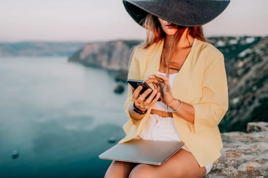 Successful business woman in yellow hat working on laptop by the sea. Pretty lady typing on computer at summer day outdoors. Freelance, travel and holidays concept.