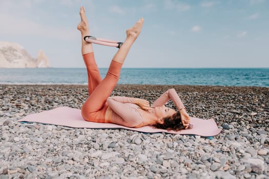 Middle aged well looking woman with black hair doing Pilates with the ring on the yoga mat near the sea on the pebble beach. Female fitness yoga concept. Healthy lifestyle, harmony and meditation.