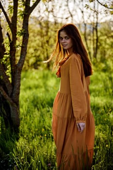 beautiful, a woman stands in a long orange dress, in the countryside, near a flowering tree, during sunset, illuminated from behind and looks sweetly into the camera. High quality photo