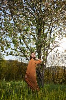 a slender, attractive woman with long red hair stands in the countryside near a flowering tree in a long orange dress and holds her hair with her hands while standing sideways to the camera. Vertical photography. High quality photo