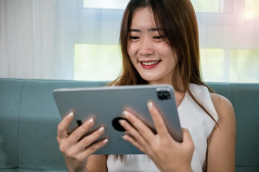 Happy Asian woman playing smart tablet at home while relaxing on sofa living room, Beautiful female sitting on couch using digital tablet pc, technology social media