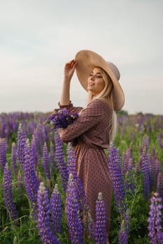 A beautiful woman in a straw hat walks in a field with purple flowers. A walk in nature in the lupin field.