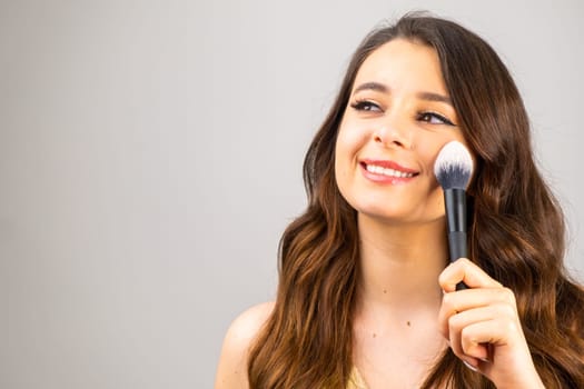 Portrait of an attractive lovely young woman holding makeup blusher brush on the grey background with copy space.