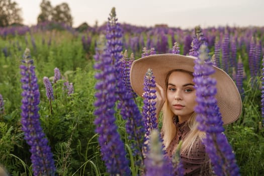 A beautiful woman in a straw hat walks in a field with purple flowers. A walk in nature in the lupin field.