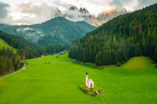 Aerial view of the Church of St. Johann against the Geisler peaks covered with clouds in Santa Maddalena village, Val Di Funes, Italy.