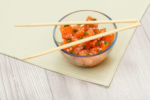 Hawaiian salmon poke with green onions and sesame seeds in glass bowl with chopsticks on kitchen napkin. Top view. Organic seafood.