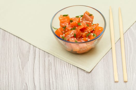 Hawaiian salmon poke with green onions and sesame seeds in glass bowl with chopsticks on kitchen napkin. Top view. Organic seafood.