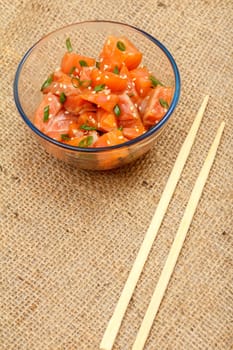 Hawaiian salmon poke with green onions and sesame seeds in glass bowl with chopsticks on sackcloth. Top view. Organic seafood.