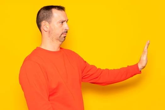 Hispanic man with a beard in his 40s wearing a red sweater gesturing stop while standing in profile, isolated over yellow background