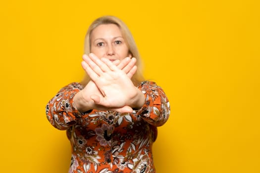 Pretty blonde Caucasian woman in her 40s wearing a patterned sweater, making the prohibition gesture with crossed palms in front of the camera, isolated over yellow studio background
