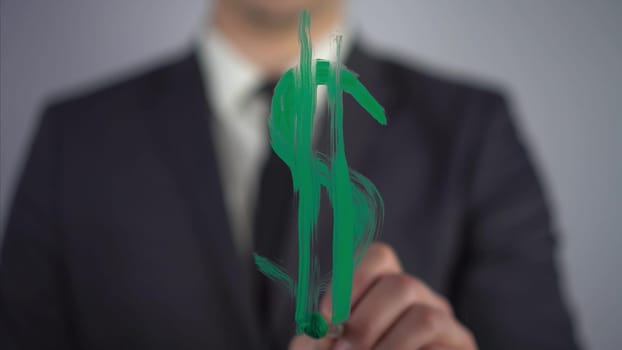 A young businessman draws a dollar symbol on a transparent glass. A man draws with green paint and a brush close-up. 4k