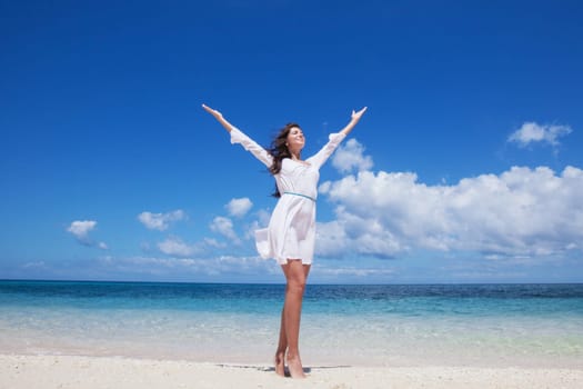 Woman in white dress posing in tropical sea beach with arms raised