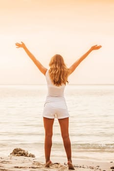 Beautiful young woman standing on sandy beach against orange sky at sunset. Summer travel. Happy slim girl in white with raised up arms on the seashore