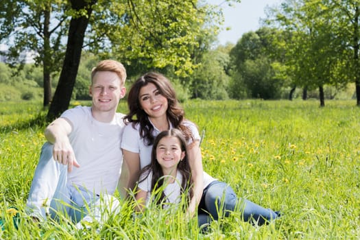 Happy smiling family of parents and daughter sitting on green grass in park