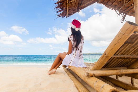 Woman in Santa hat sitting at chaise lounge with straw parasol on white sandy beach at Philippines