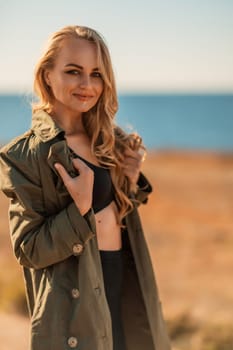 Portrait blonde sea cape. A calm young blonde in an unbuttoned khaki raincoat stands on the seashore, under the raincoat there is a black skirt and top.