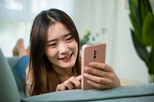 Asian young woman using smart mobile phone lying on sofa in living room room at home, Smiling attractive female relaxed reading text message on smartphone