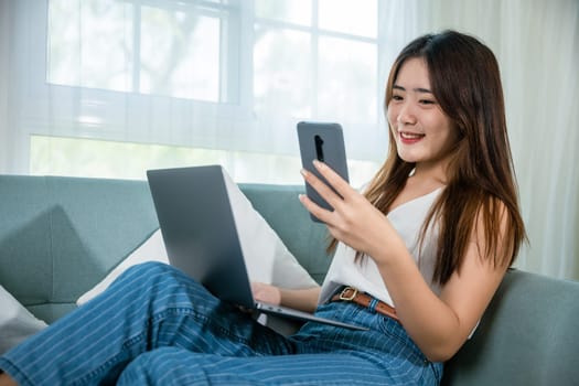 Asian young woman using smart mobile phone while sitting on sofa in living room at home with laptop computer, Happy female use netbook browsing and payment shopping online with smartphone