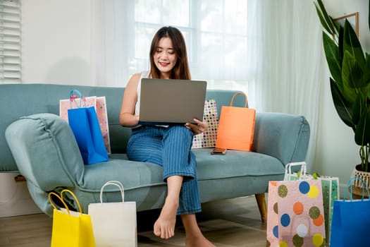 Asian young woman sitting on sofa with shopping bags using laptop computer to online shopping, Happy female satisfied customer with purchase on web site in living room at home