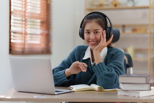 Online education, e-learning. Asian woman in stylish casual clothes, studying using a laptop, listening to online lecture, taking notes, online study at home.