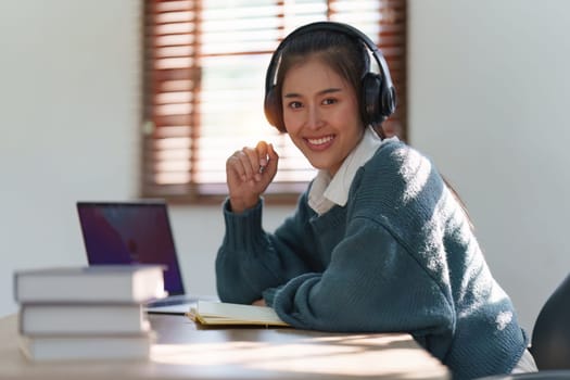 Online education, e-learning. Asian woman in stylish casual clothes, studying using a laptop, listening to online lecture, taking notes, online study at home.