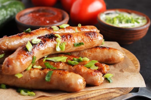 Delicious sausages on a wooden board with various sauces and fresh vegetables on a wooden table.