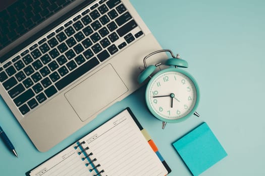 Laptop with an alarm clock, a diary notebook and others work and educational materials on blue background for the concept of work, study and time management.