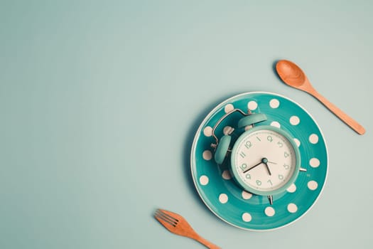 An alarm clock on an empty plate and cutlery set against blue background for the concept of food, time management, losing weight and eating on time.