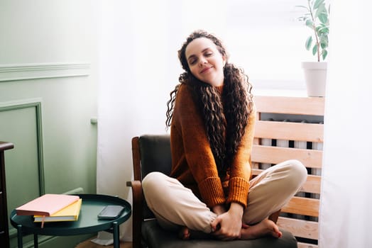 Young relaxed smiling pretty woman relaxing sitting on chair at home. Happy positive beautiful lady feeling joy enjoying wellbeing and lounge chilling near window in modern cozy apartment interior