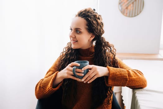 Young smiling pretty woman holding cup drinking warm tea or coffee relaxing dreaming at home. Happy positive calm lady enjoying hot drink with mug in hands daydreaming in cold cozy winter morning