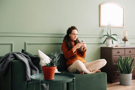 Young woman using mobile cell phone sitting on the couch at home. A pretty relaxed lady holding a smartphone buying in an e-commerce shop watching videos online, texting, or checking social media news