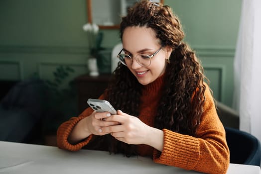 Young smiling woman wearing glasses holding a smartphone using cellphone modern technology, looking at mobile, checking cell phone apps, texting, and browsing the internet for shopping sitting at home