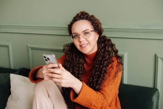 Smiling young pretty woman sitting at home using apps on cell phone technology, happy lady holding smartphone, laughing, looking at camera, relaxing with cellphone in hands checking cellular device