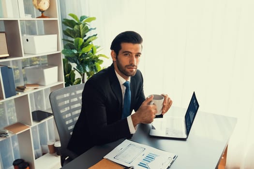 Modern professional businessman at modern office desk using laptop to work with coffee in his hand. Diligent office worker working on computer notebook in his office work space. fervent