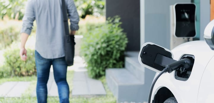 Focus electric car charging at home charging station with blurred progressive man walking in the background. Electric car using renewable clean for eco-friendly concept.