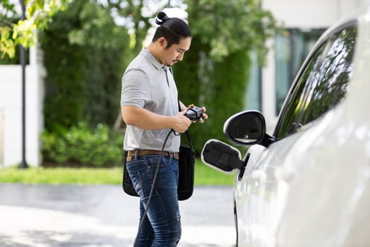 Progressive asian man install cable plug to his electric car with home charging station in the backyard. Concept use of electric vehicles in a progressive lifestyle contributes to clean environment.