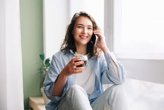 Adorable cute funny woman with a mug of black tea talking on the phone sitting on the windowsill in the kitchen, having fun, and enjoying pleasant conversation with her sister or best friend.