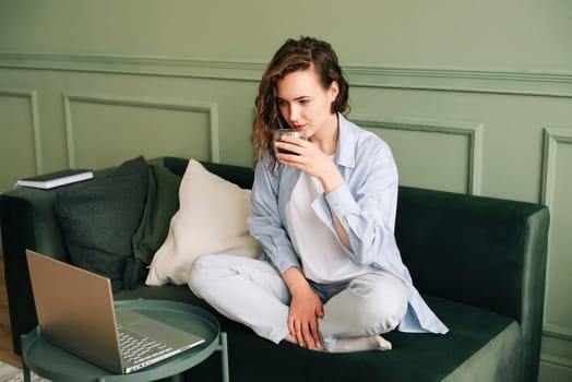 Adorable female drinking hot tea sitting on a sofa near a big window and watching webinars and online tutorials on a laptop using a wireless connection, studying from home.