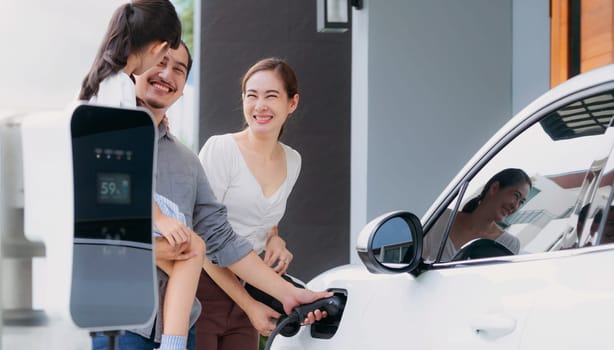 Progressive young parents and daughter with electric vehicle and home charging station. Green and clean energy from electric vehicles for healthy environment. Eco power from renewable source at home.