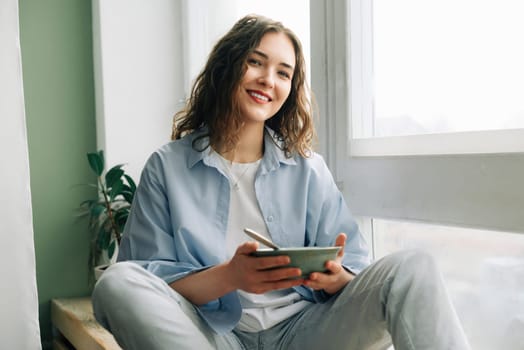 A young woman in jeans and a shirt sitting on the window sill looking through the big window the city holding a bowl of breakfast in her hands, smiling noticing something funny outside.