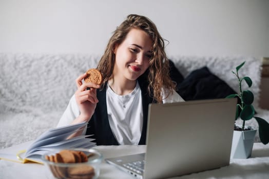 Young woman watching series or podcasts looking at the screen with interest holding cookies in hand sitting at the kitchen table with a glass jar and lapto, smiling happily Unconscious eating disorder