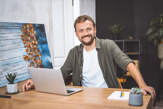 Concept of remote work, freelance, and productivity in digital age. Cheerful and productive man, works from home, He smiling, focusing on his work, surrounded by digital technology and equipment. . High quality photo