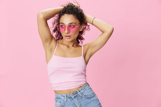 Happy woman hands up waxing her armpits, shaving, with curly hair in a pink tank top and jeans on a pink background wearing sunglasses with a nice tan, copy space. High quality photo