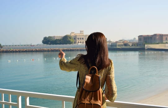 A woman in a long dress with a backpack indicates the direction with her hand to the sea. Back view. Travel concept